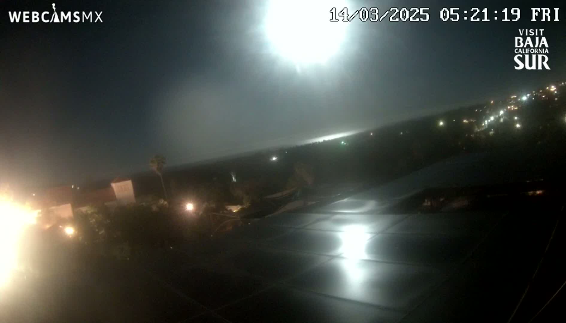 A nighttime, hazy view from an elevated vantage point shows a bright light in the distance over a dark landscape with scattered lights, and a dark, reflective surface in the foreground.