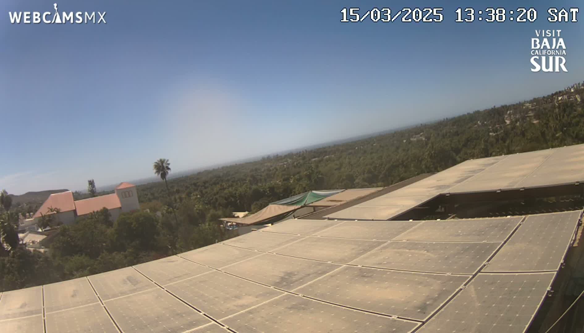 A large solar panel array on a building overlooks a sunny, tree-covered landscape extending to a distant ocean under a clear blue sky.