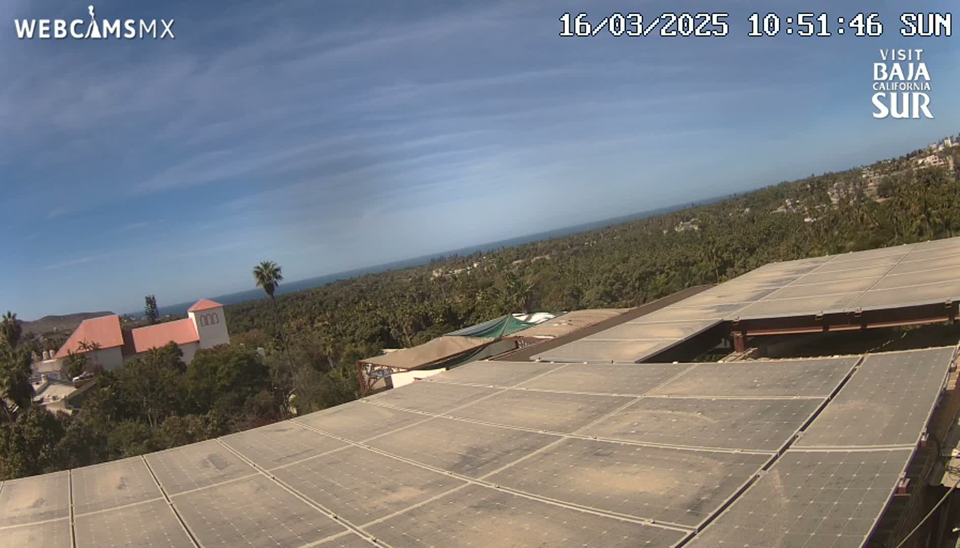 A large solar panel array sits atop a building overlooking a sunny coastal town and ocean under a clear blue sky.