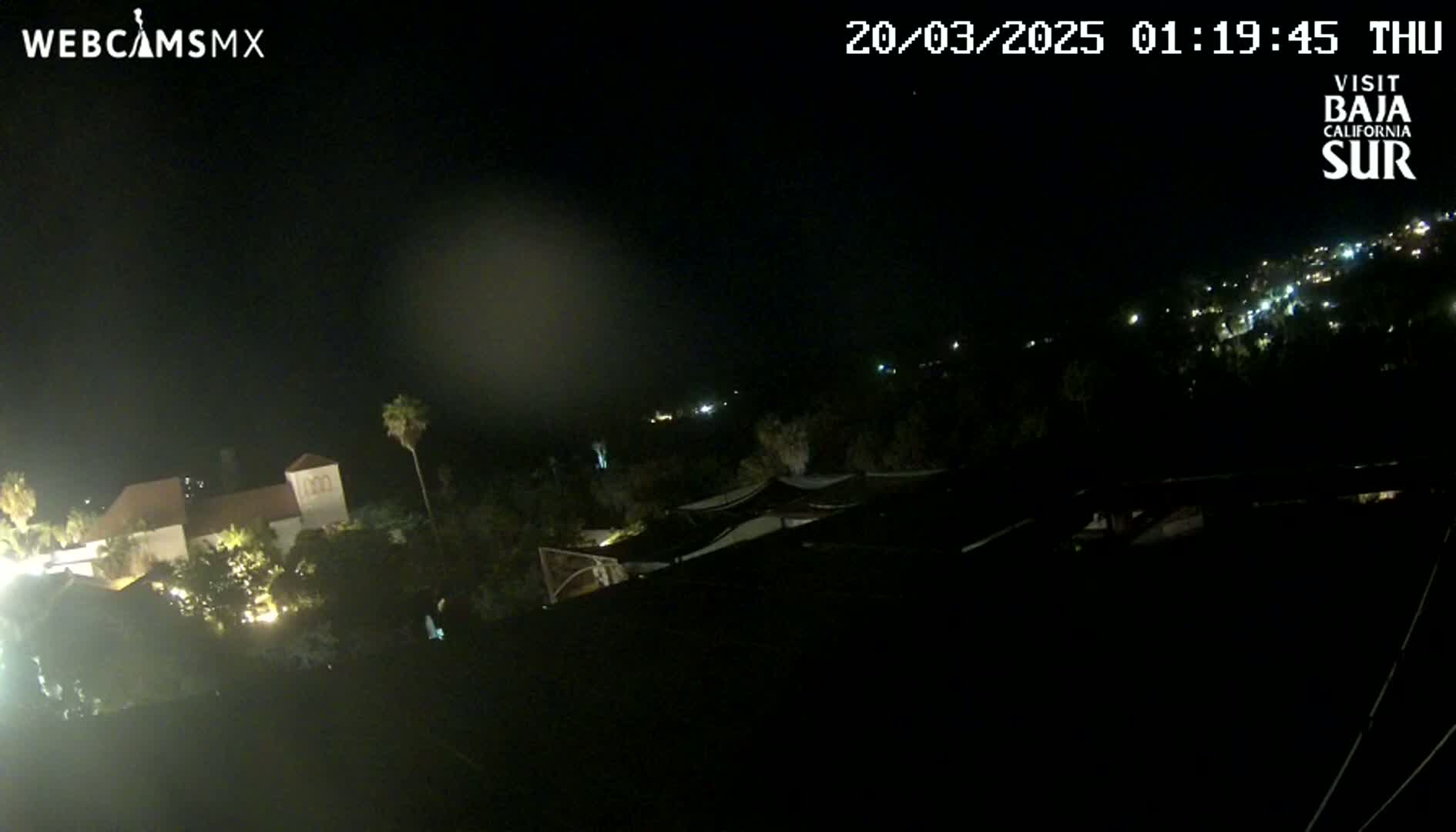 A nighttime view shows a building with lights on, surrounded by dark vegetation and a distant hillside with scattered lights under a clear, dark sky.