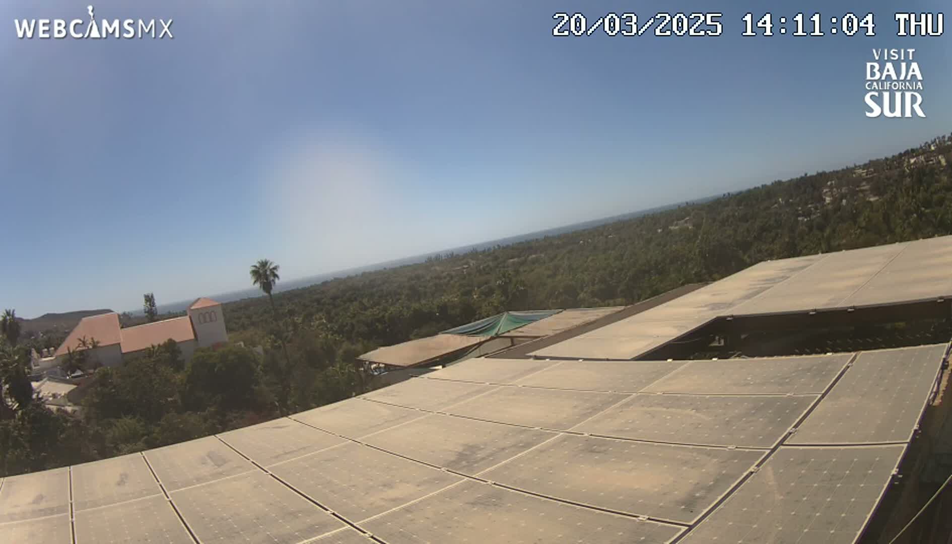 A large solar panel array sits atop a building overlooking a sunny landscape with a distant ocean view and some buildings nestled among green trees.