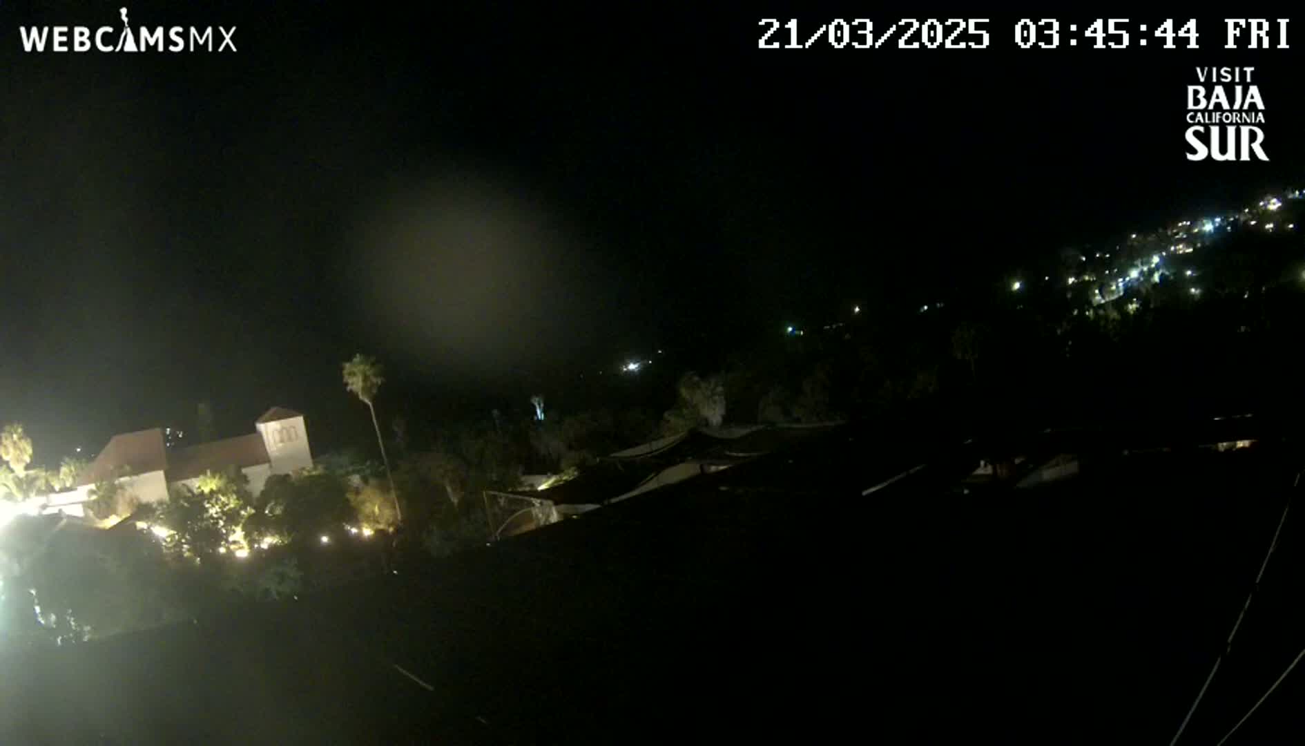 A nighttime, high-angle view shows a building complex with sparse outdoor lighting, palm trees, and a distant town illuminated under a dark sky.