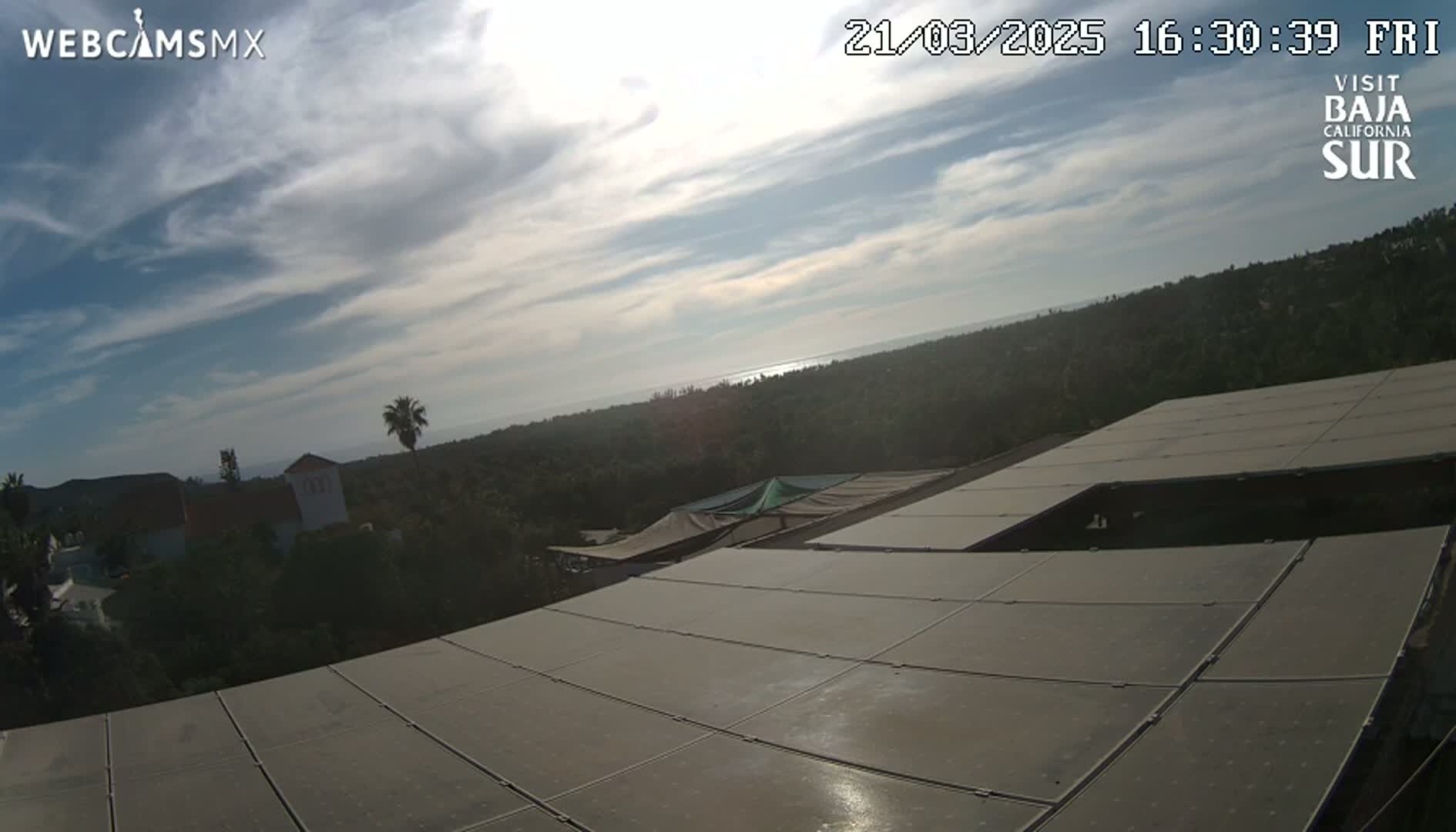 A partly cloudy sky overlooks a landscape of vegetation and buildings, with a large solar panel array in the foreground.