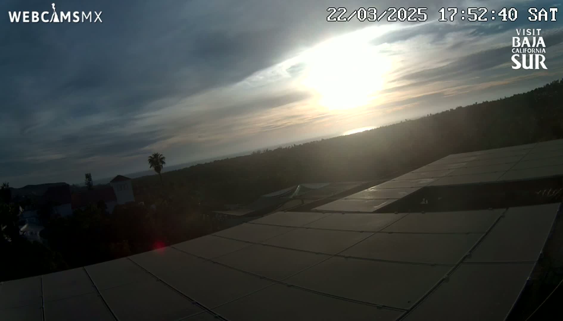 A partly cloudy sunset over a hilly landscape with a large solar panel array in the foreground.
