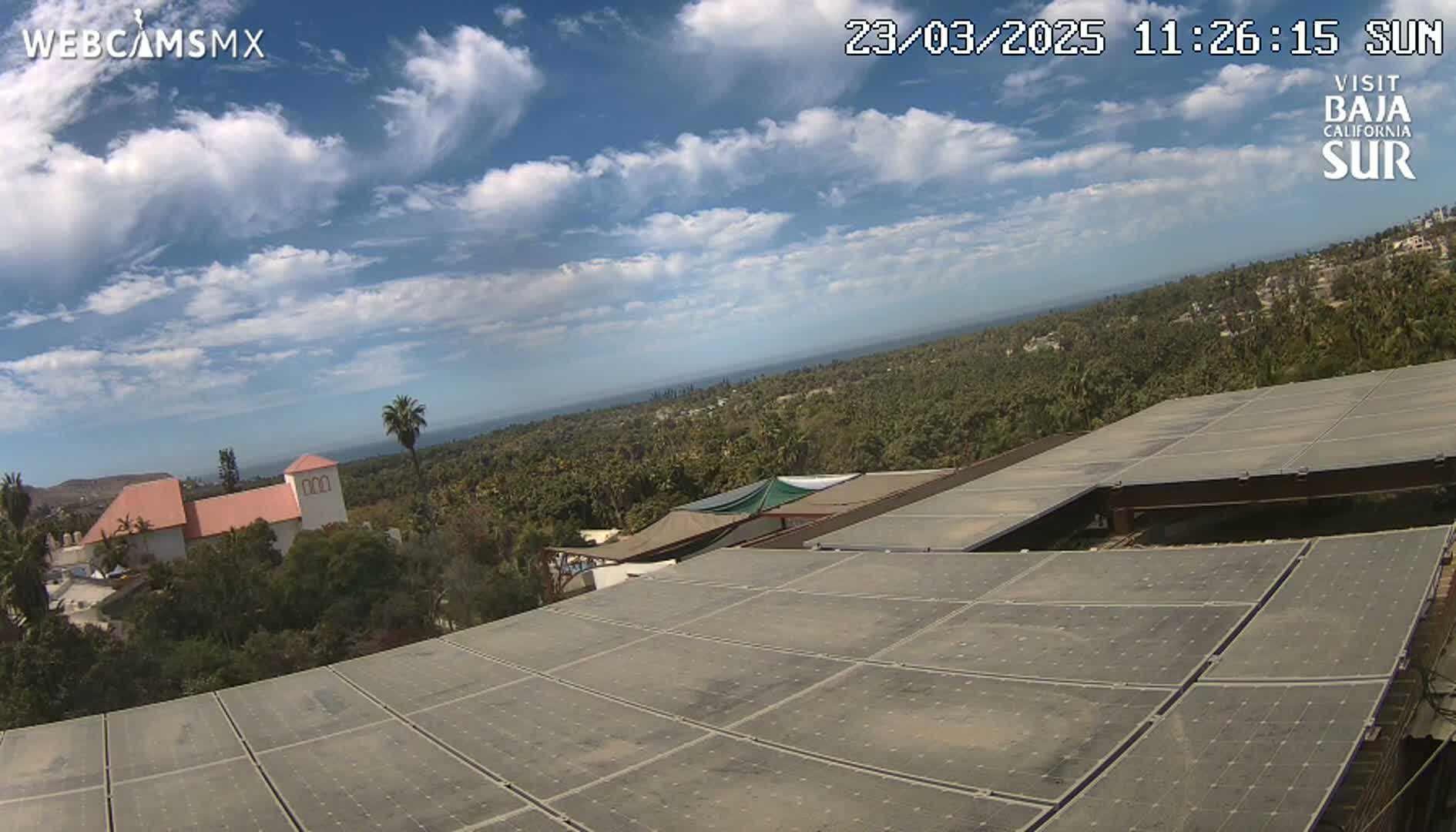 A large solar panel array sits atop a building overlooking a sun-dappled ocean vista and a landscape of lush green vegetation under a partly cloudy sky.
