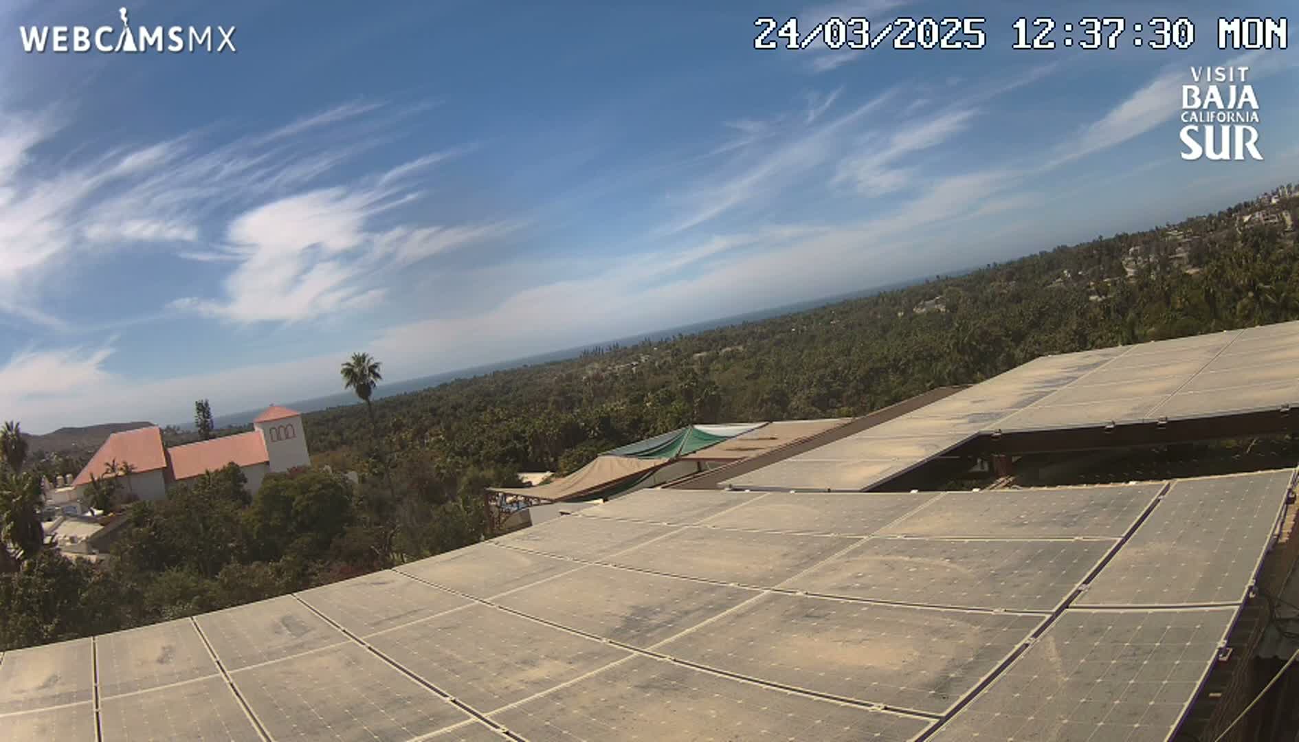 A mostly sunny day reveals a large expanse of solar panels atop a building overlooking a coastal town nestled amongst lush green vegetation and a calm ocean.