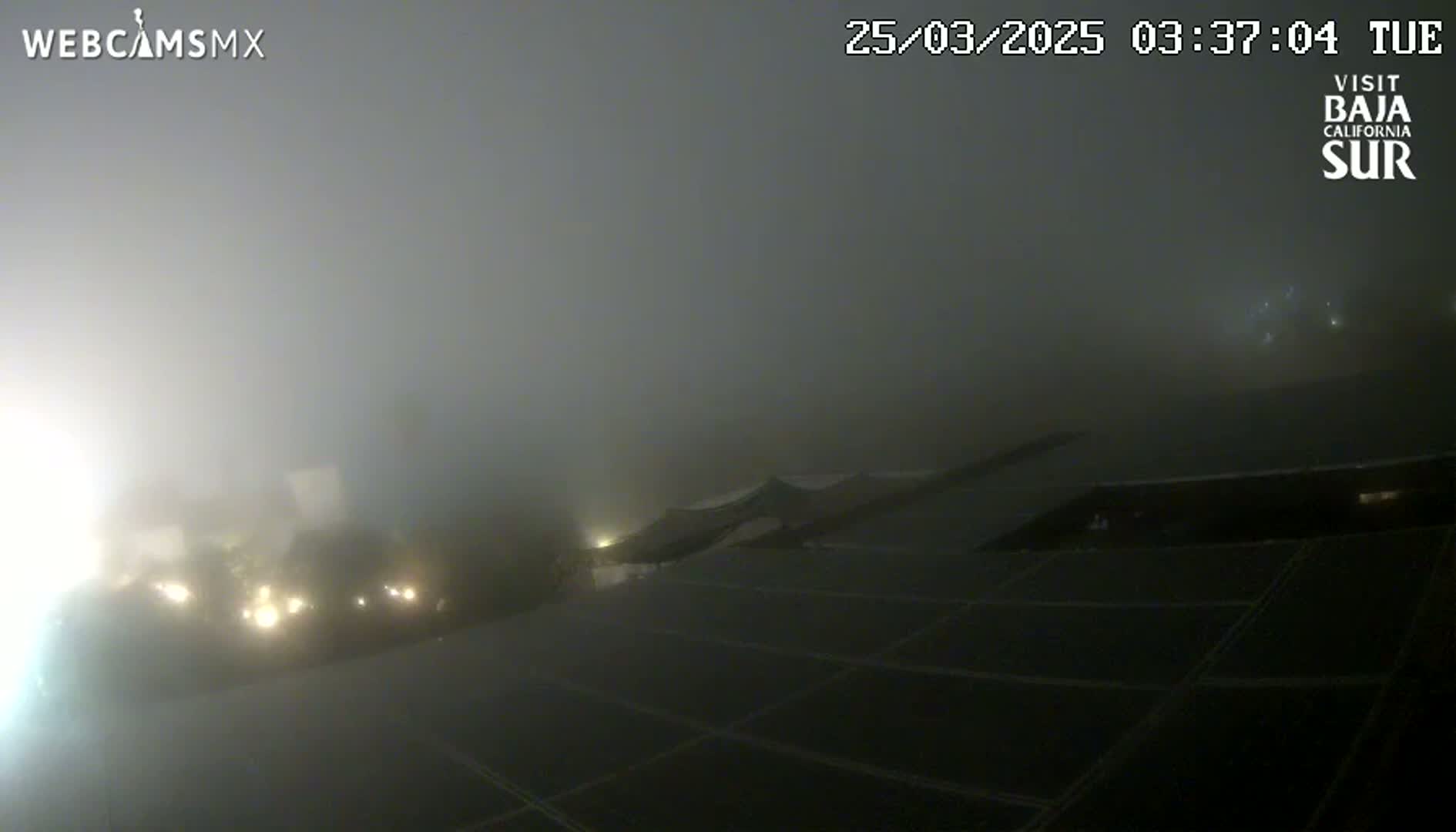 A thick fog obscures the view of a dark, flat surface divided into sections, with faint lights visible in the distance.