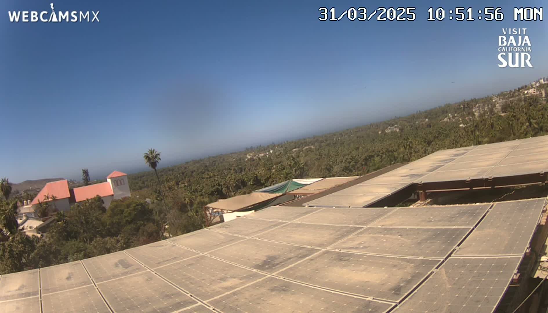 A large solar panel array sits atop a building overlooking a lush, green landscape under a clear blue sky.