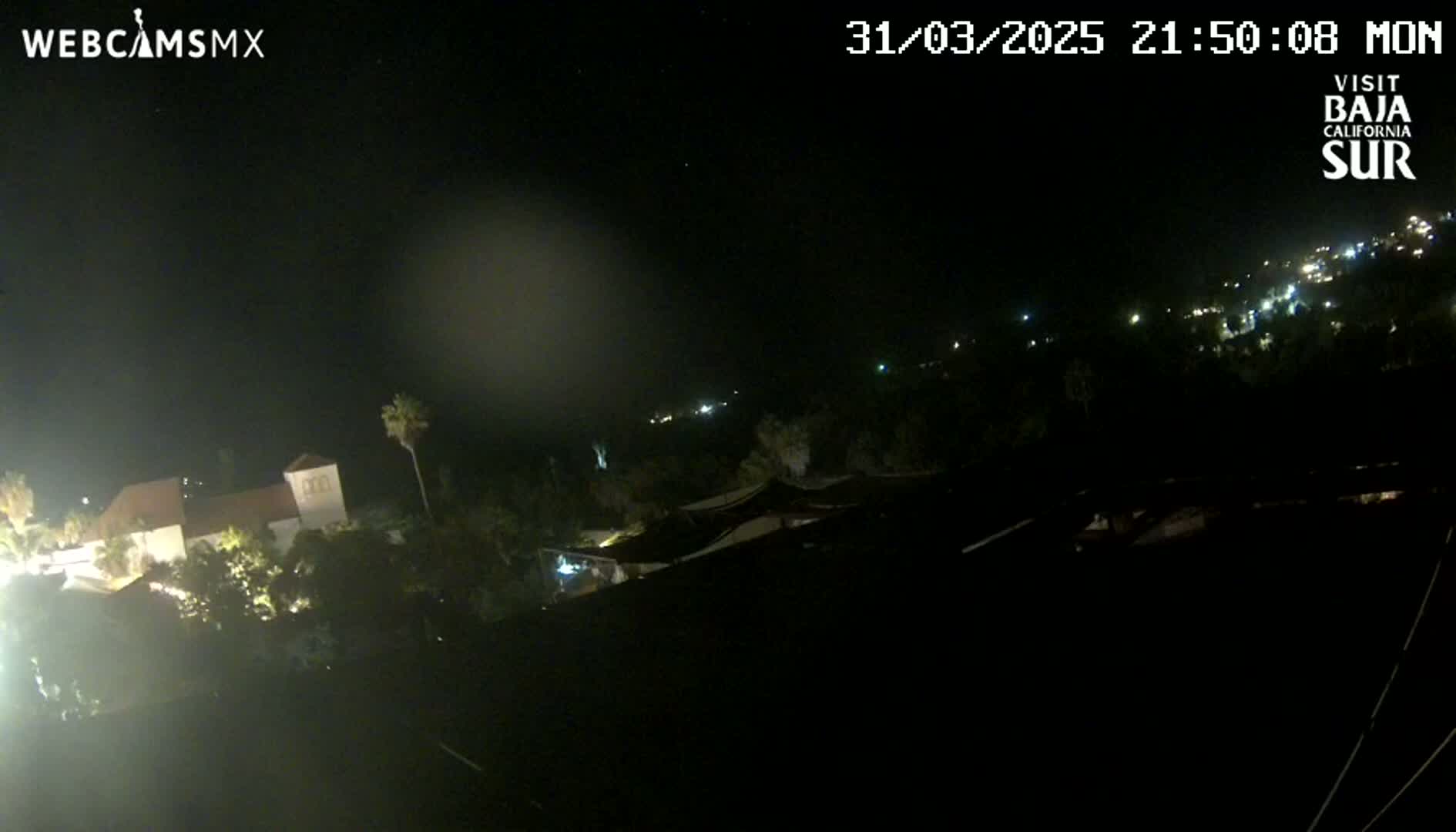 A nighttime view shows a building complex partially illuminated, surrounded by dark vegetation, and overlooking a hillside with scattered lights under a clear, dark sky.