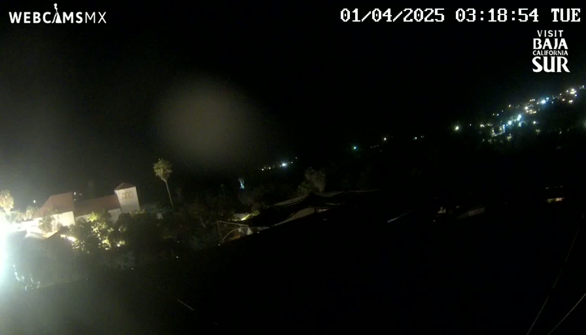 A nighttime, long shot shows a building with lights on and some vegetation, with a hazy sky and distant lights in the background.