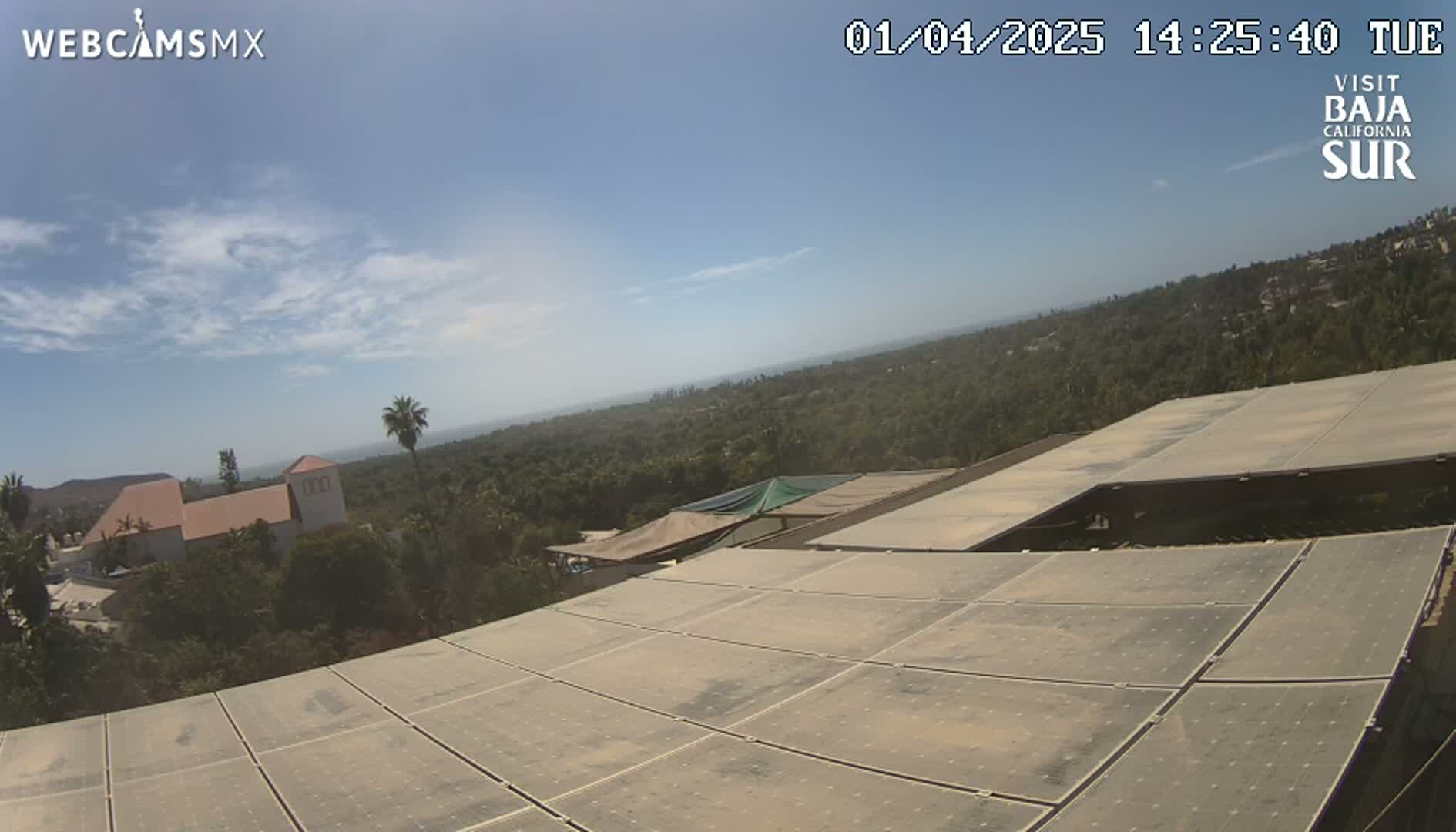 A view from a dusty rooftop shows a partly sunny sky over a landscape of green vegetation, buildings, and a distant ocean under a mostly clear sky.