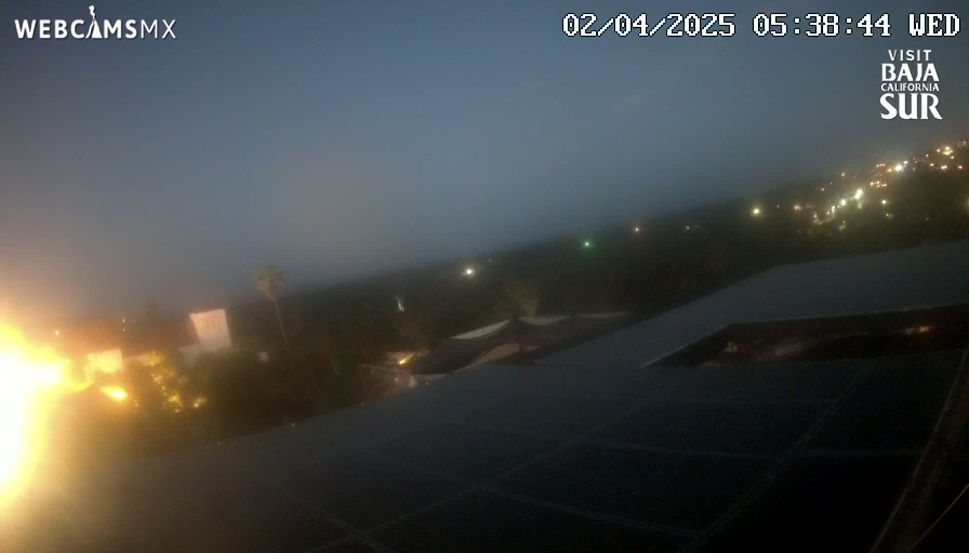A foggy, twilight view shows a rooftop solar panel array overlooking a dimly lit residential area.