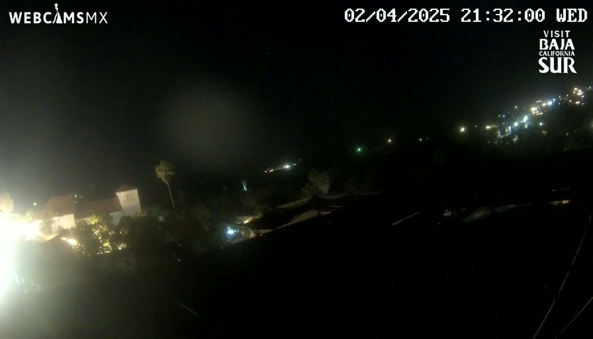 A nighttime view shows a dimly lit town with buildings and trees, under a dark, hazy sky.