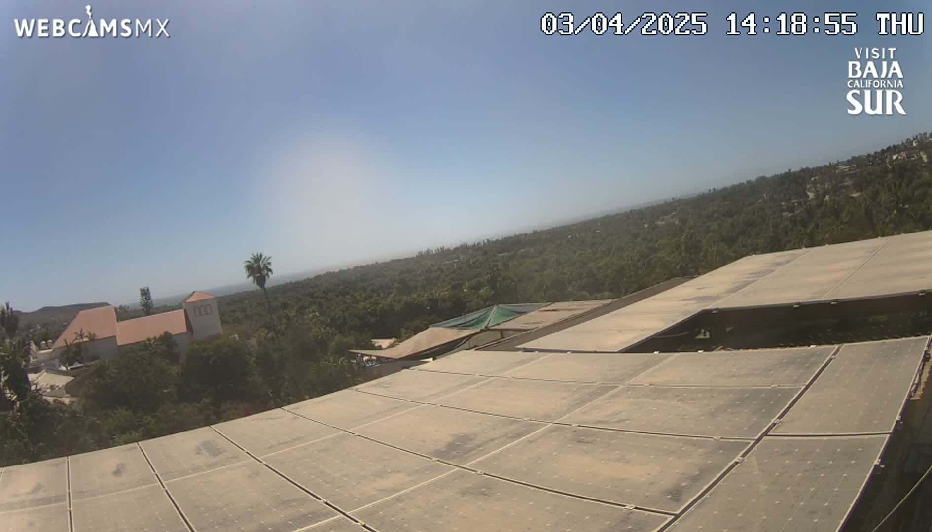 A large array of solar panels sits atop a building overlooking a sunny landscape of trees, buildings, and a distant ocean under a clear blue sky.