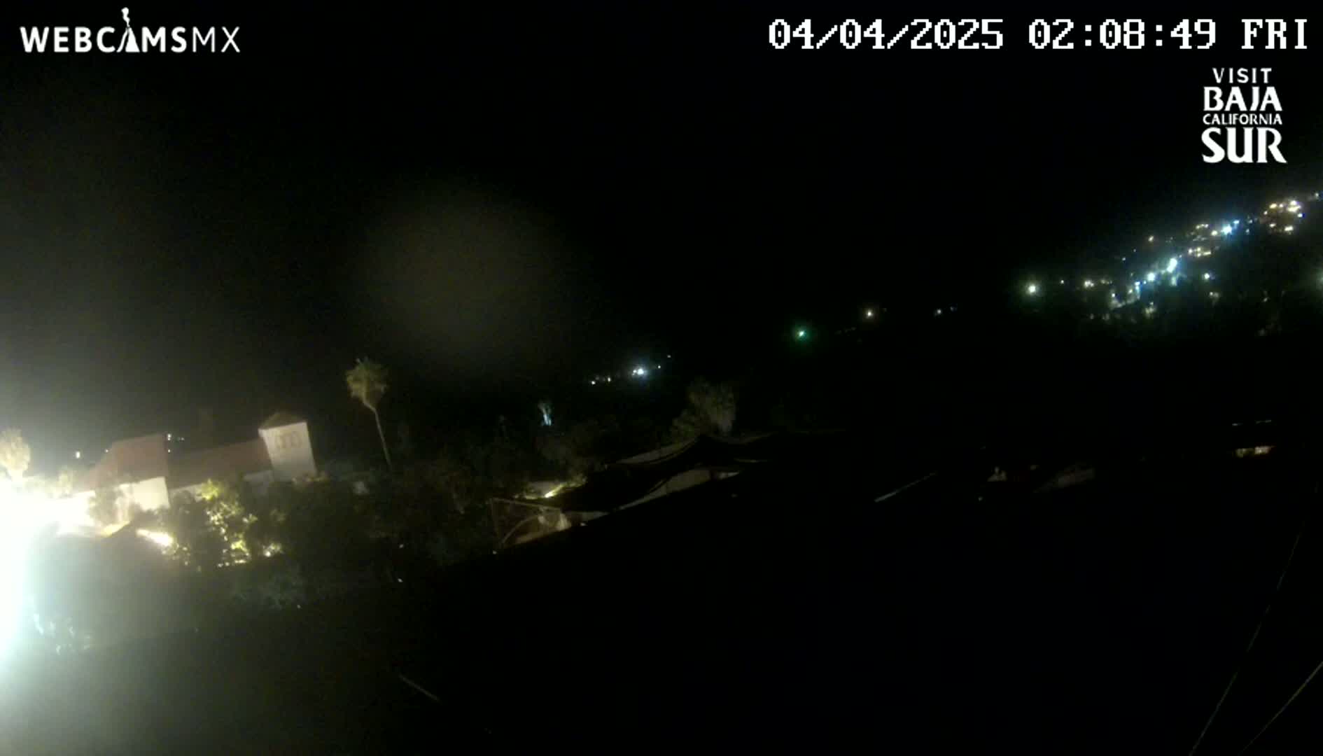 A nighttime, high-angle view shows a dark landscape with a few lights visible in the distance and some structures illuminated closer to the camera.