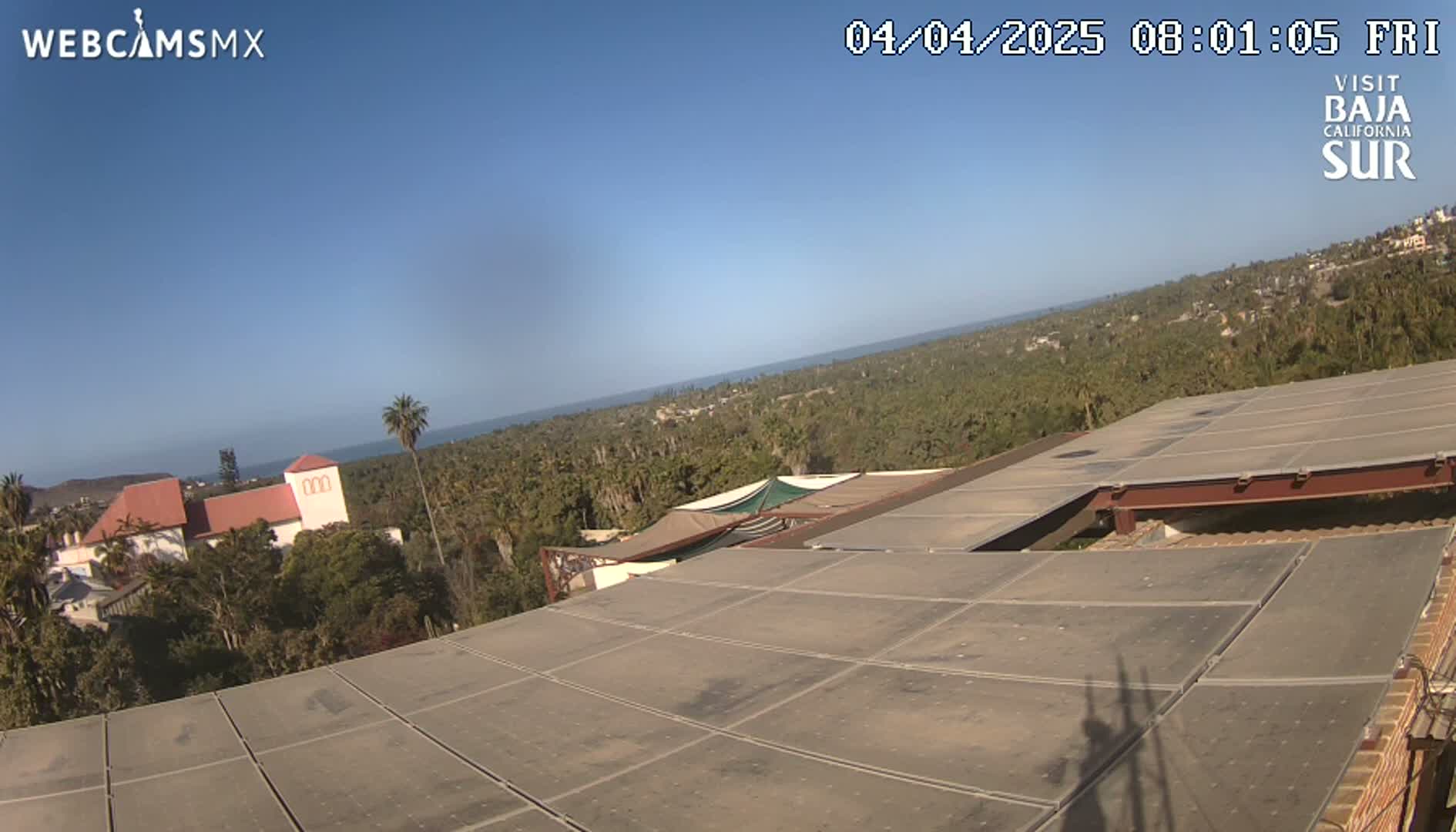 A flat rooftop with a sectioned surface overlooks a clear blue ocean and a sprawling landscape of green vegetation under a bright, sunny sky.