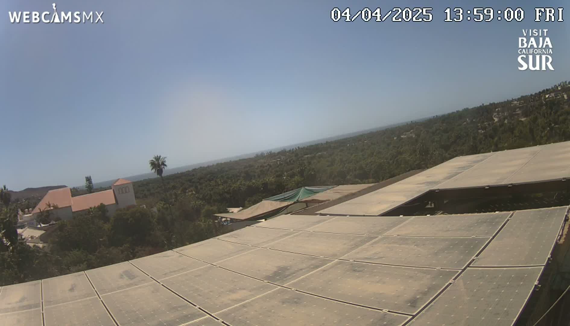 A large solar panel array sits atop a building overlooking a verdant landscape and a distant ocean under a clear, sunny sky.