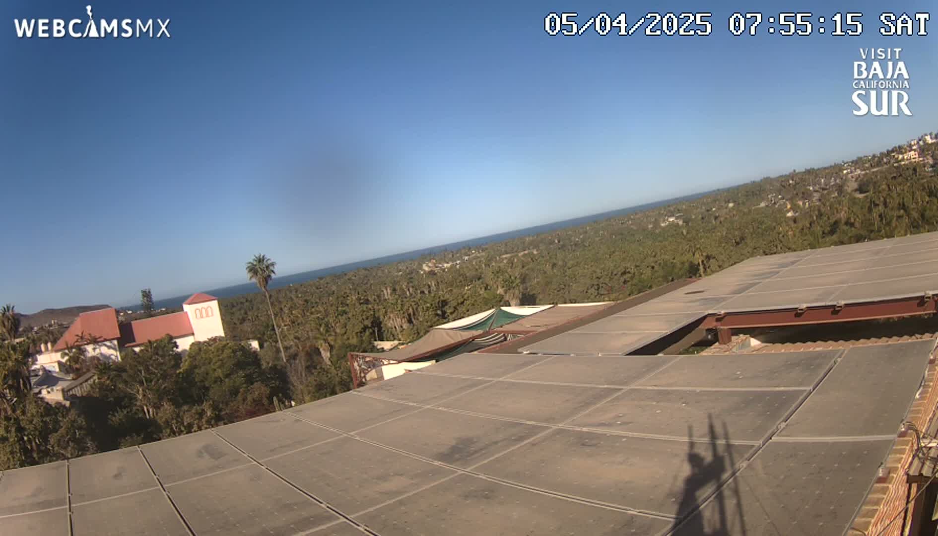 A clear, sunny day reveals a panoramic view of a coastal town nestled amongst palm trees, overlooking a calm ocean from a rooftop with a large, flat surface.