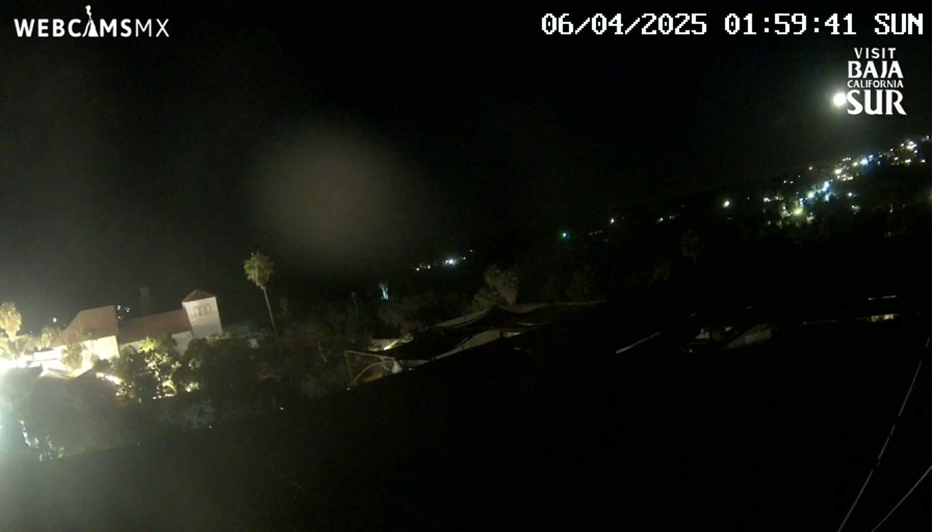 A nighttime, long shot of a building complex surrounded by trees, with distant lights visible in the background under a clear sky.
