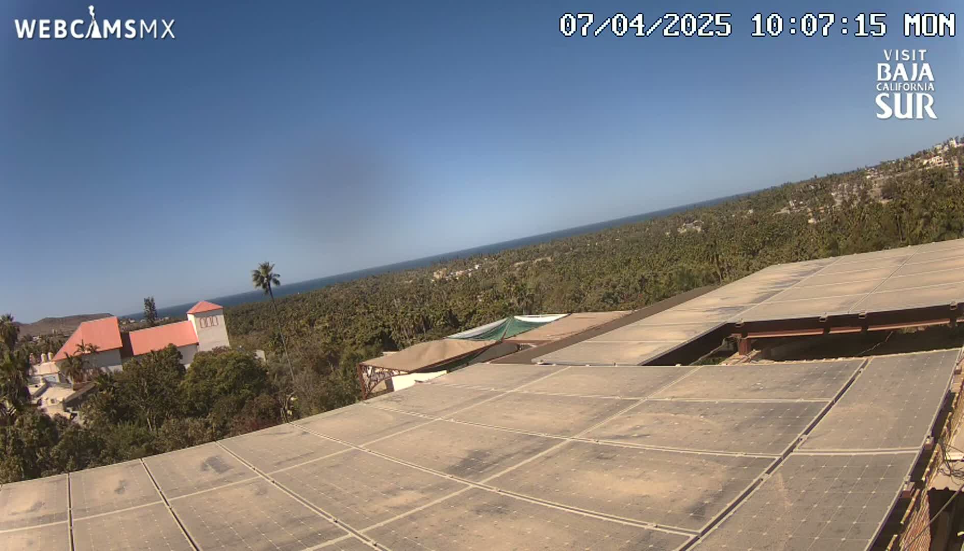 A large array of solar panels sits atop a building overlooking a coastline, under a clear blue sky.