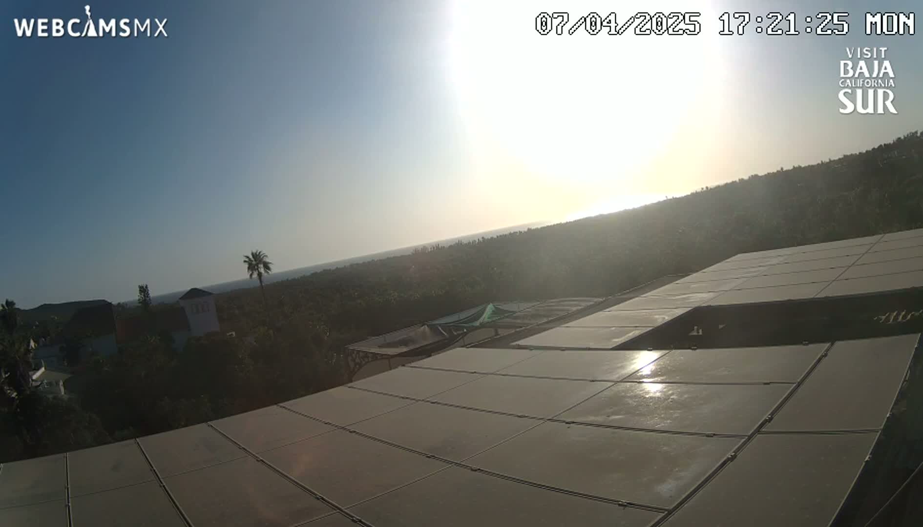 A sunlit view overlooking a partly shaded hillside with buildings and a large, flat, solar panel array, under a clear sky.