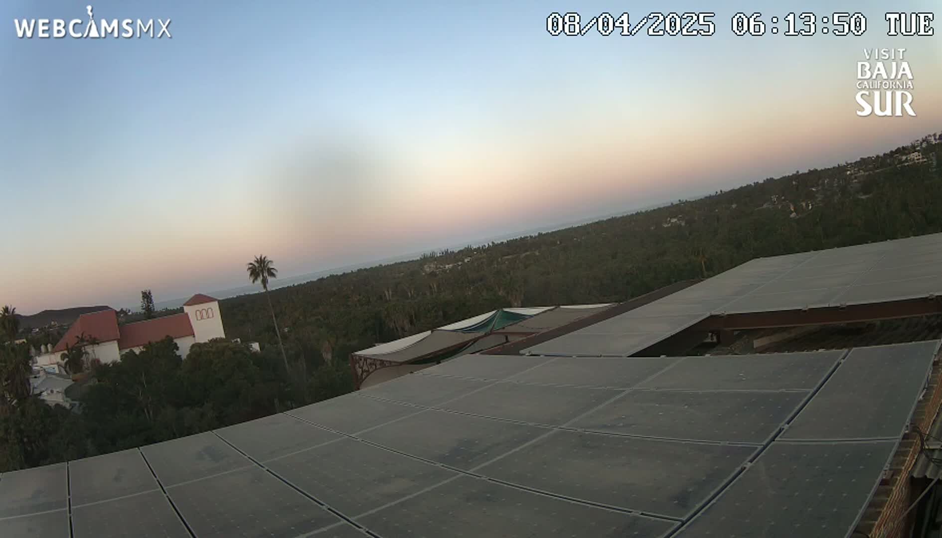 A large solar panel array on a rooftop overlooks a coastal town at sunset under a clear sky.