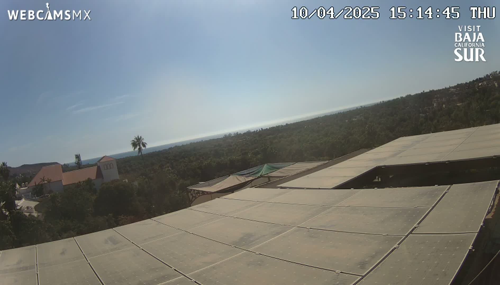 A view from a rooftop of solar panels overlooks a coastal landscape under a clear, sunny sky.