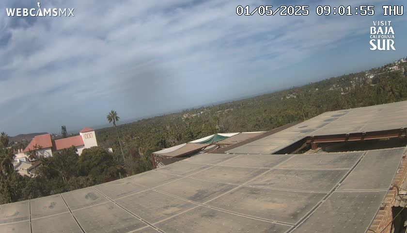 A mostly clear sky overlooks a large flat roof with a view of palm trees, buildings, and distant hills.