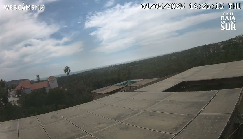 A partly cloudy day reveals a sprawling landscape of vegetation and buildings, viewed from a high vantage point overlooking a large flat rooftop.