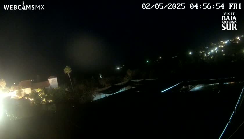 A nighttime, high-angle view shows a collection of buildings with lights illuminated, some vegetation, and a dark sky.