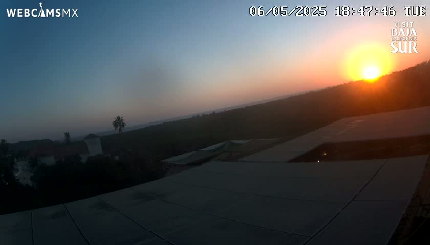 A sunset over a hilly landscape with buildings and vegetation, viewed from a rooftop.
