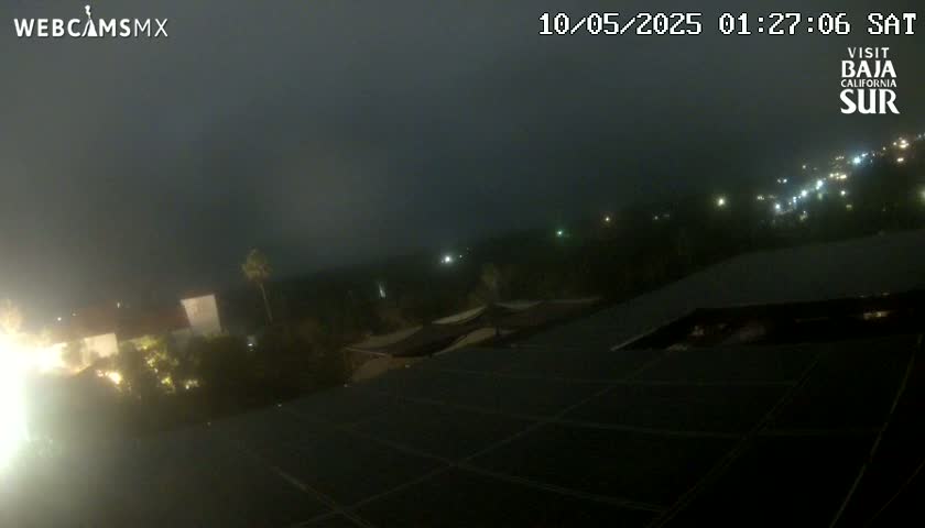 A nighttime view of a dark, hazy landscape with distant lights and a solar panel array in the foreground.