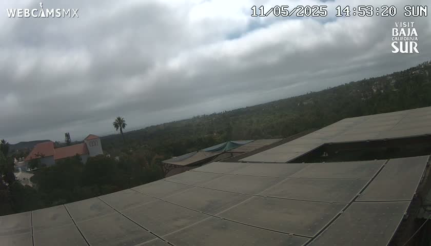 An overcast sky overlooks a hilly landscape with buildings and vegetation, partially viewed from above a large, flat, paneled surface.