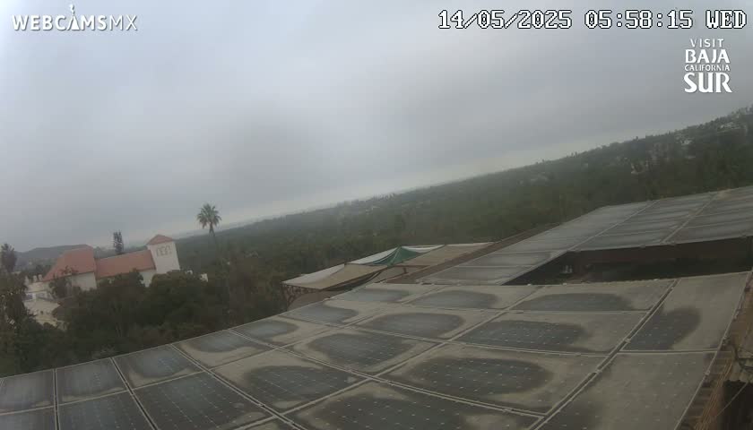 A view from a rooftop shows a cloudy sky over a hilly, wooded area with buildings and solar panels in the foreground.