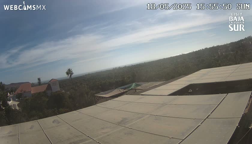 A mostly sunny day overlooks a sprawling landscape of green vegetation and buildings, viewed from a vantage point atop a structure with a large, flat, light-tan roof.