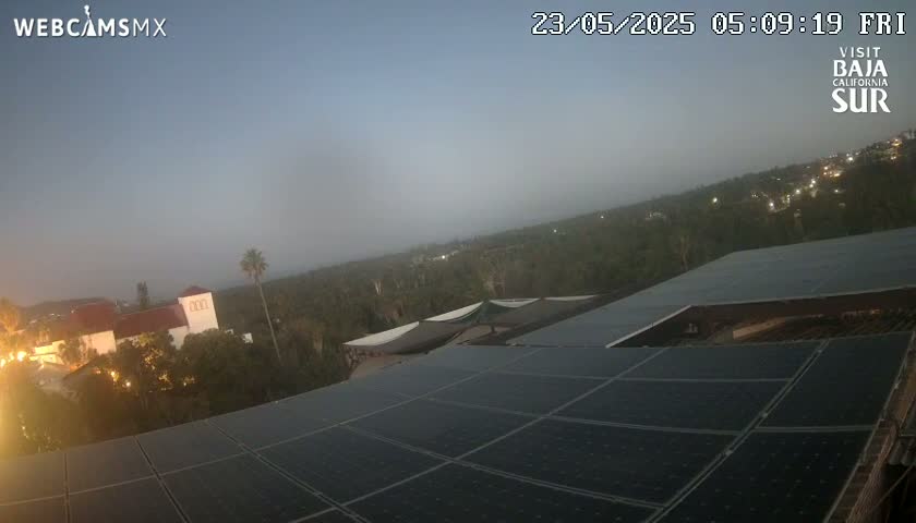 A hazy dusk scene shows a large solar panel array on a rooftop overlooking a town nestled among trees.