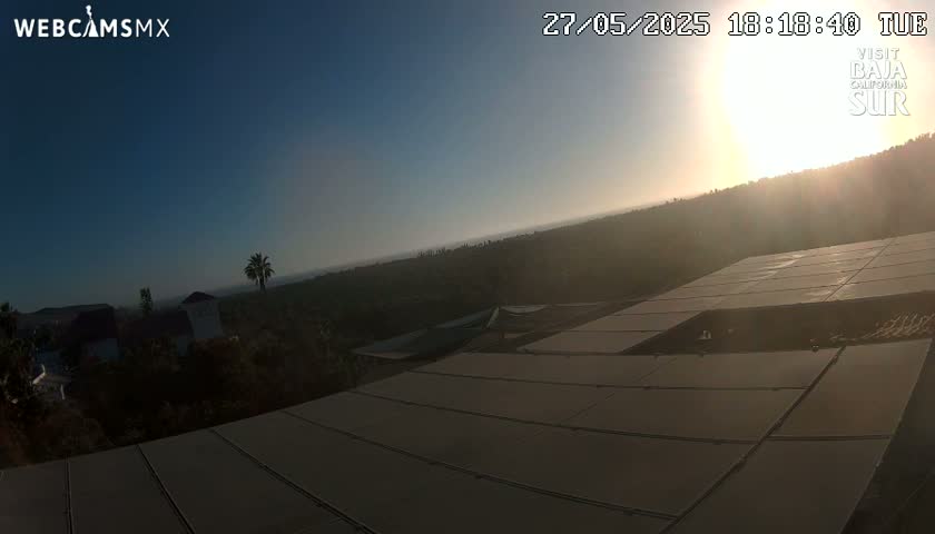 A sunlit view from a rooftop shows a hillside covered in vegetation, a few buildings in the distance, and a large solar panel array in the foreground under a clear sky.