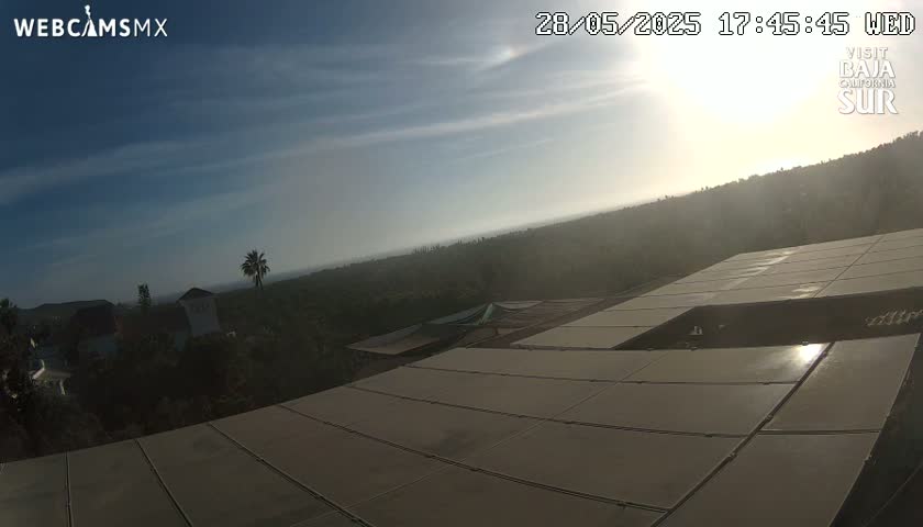 A hazy, sunny day overlooks a hilly landscape with buildings and vegetation, seen from a rooftop covered in solar panels.