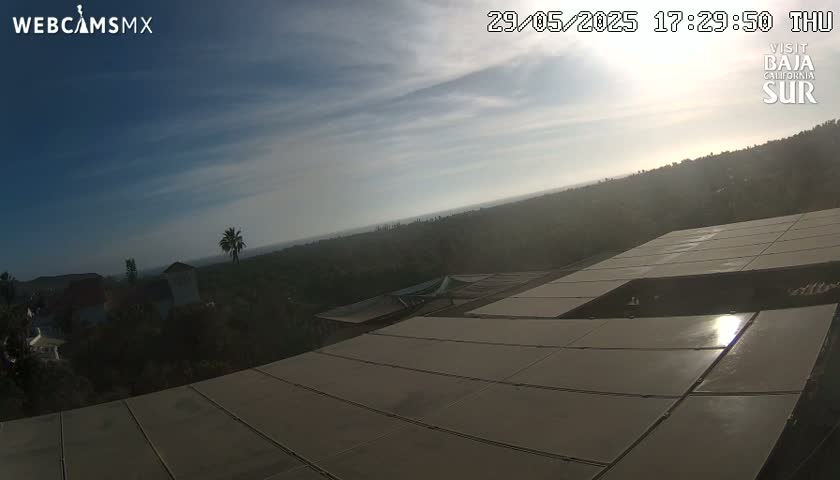 A mostly clear, sunny sky overlooks a hilly landscape with houses and a large solar panel array on a roof.