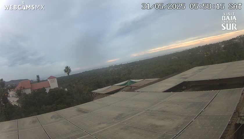 An overcast sky reveals a sunset horizon over a landscape of lush vegetation, buildings, and a large flat rooftop surface.