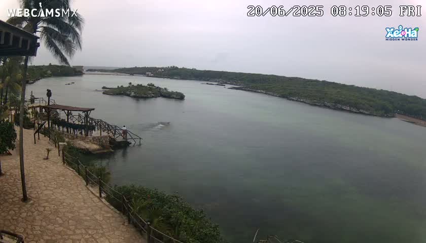 An overcast day reveals a tranquil cove with calm, greenish water, surrounded by lush green vegetation and a stone pathway leading to a small wooden structure near the water's edge.