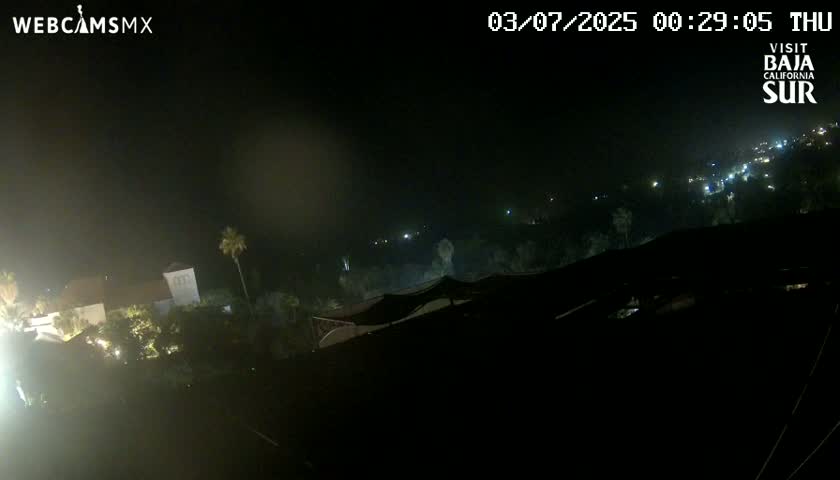 A nighttime, aerial view shows a partially lit landscape with buildings and vegetation under a dark sky.