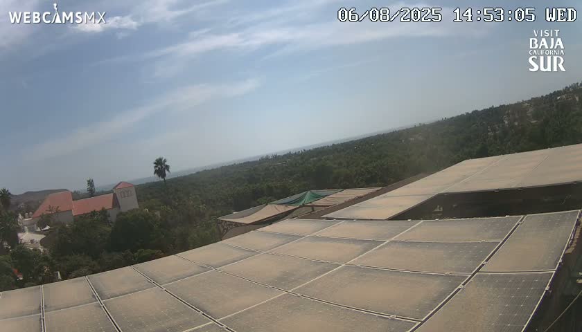A partly cloudy sky overlooks a dusty array of solar panels on a building, with a distant view of buildings and green landscape under a bright sun.