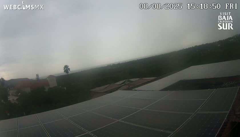 A partly cloudy sky overlooks a dusty array of solar panels on a building, with a distant view of buildings and green landscape under a bright sun.
