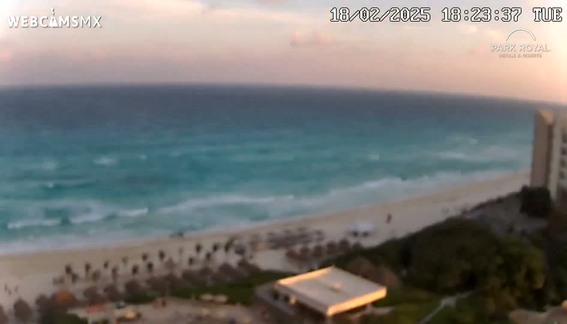 A beach with turquoise water and small waves under a partly cloudy, pinkish sunset sky is viewed from a high vantage point, showing beach umbrellas and a building in the foreground.