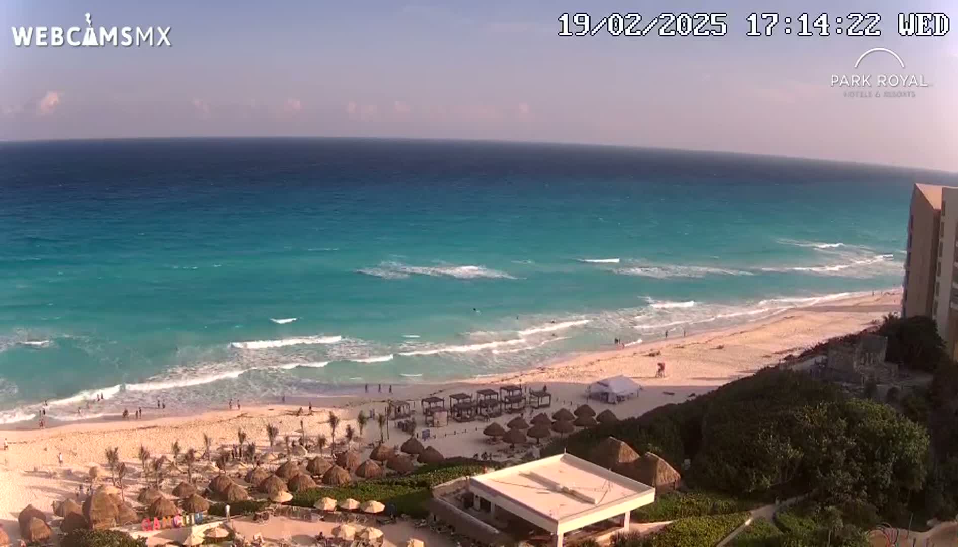 A beach with turquoise water and small waves under a mostly clear sky is seen from an elevated vantage point, showing resort buildings and beach umbrellas in the foreground.