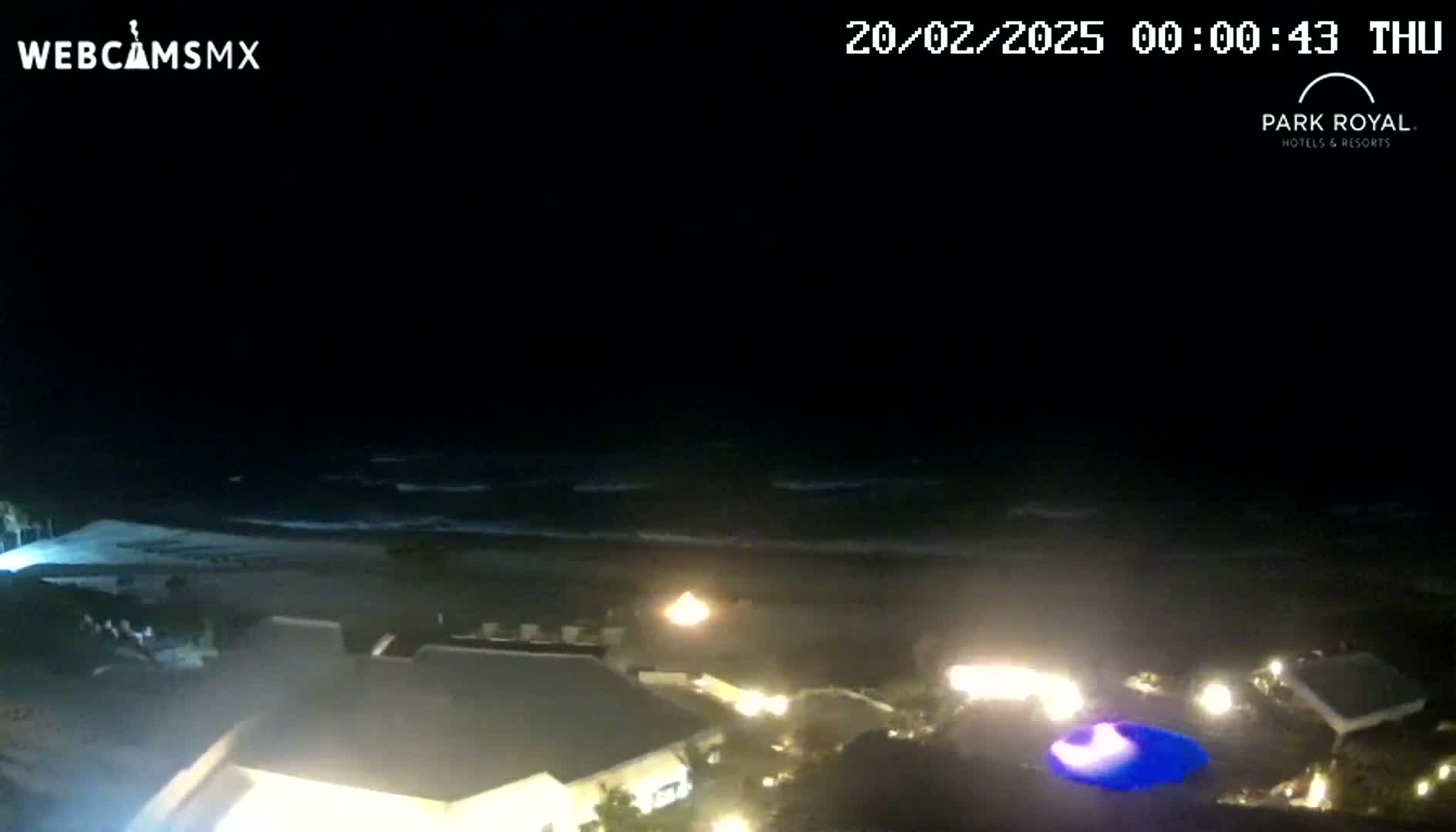 A nighttime coastal scene shows a dark ocean with waves gently lapping the shore near a dimly lit resort.