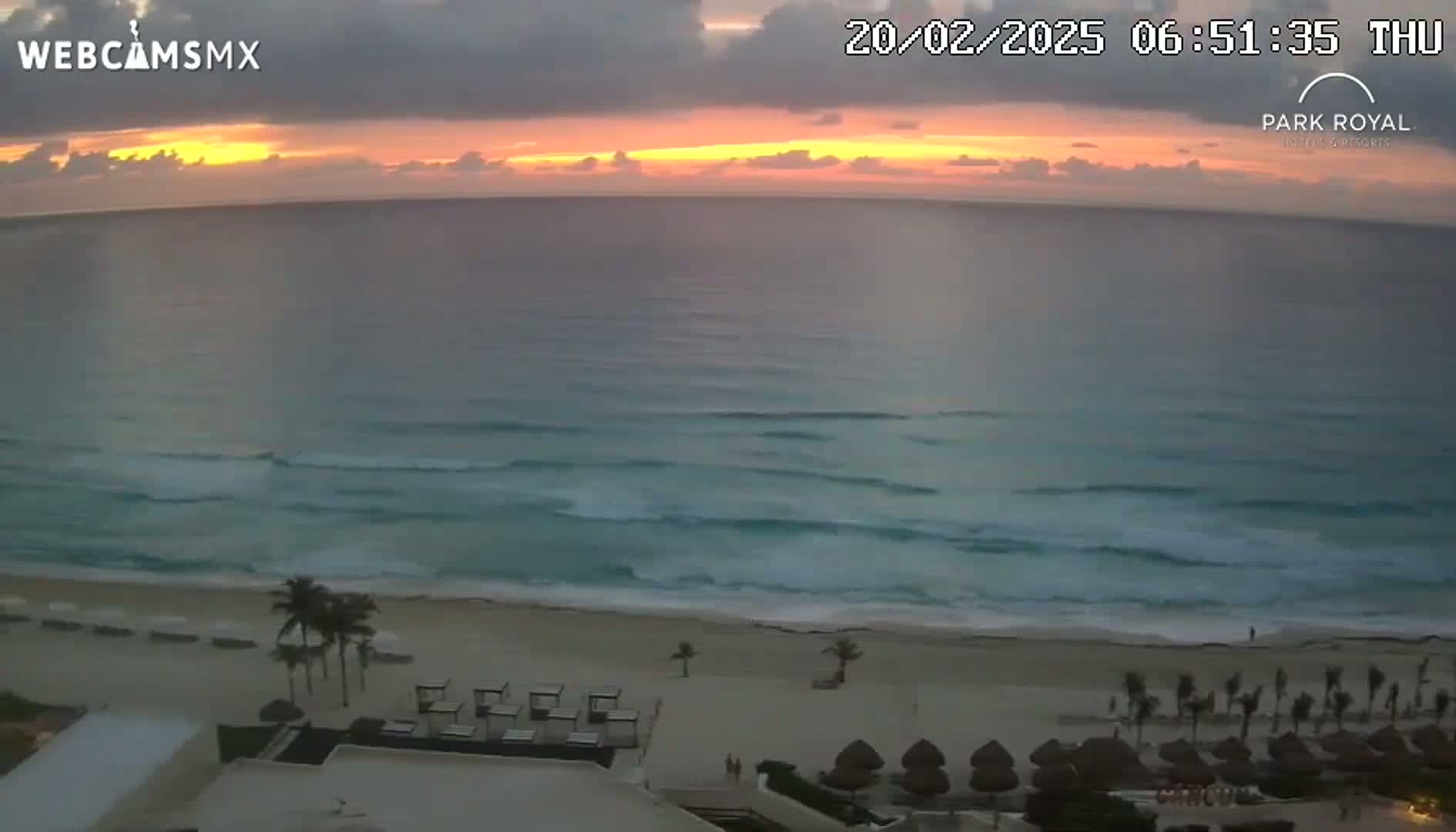 A sunrise over a beach with calm waves, showing a resort with palm trees and beach umbrellas.