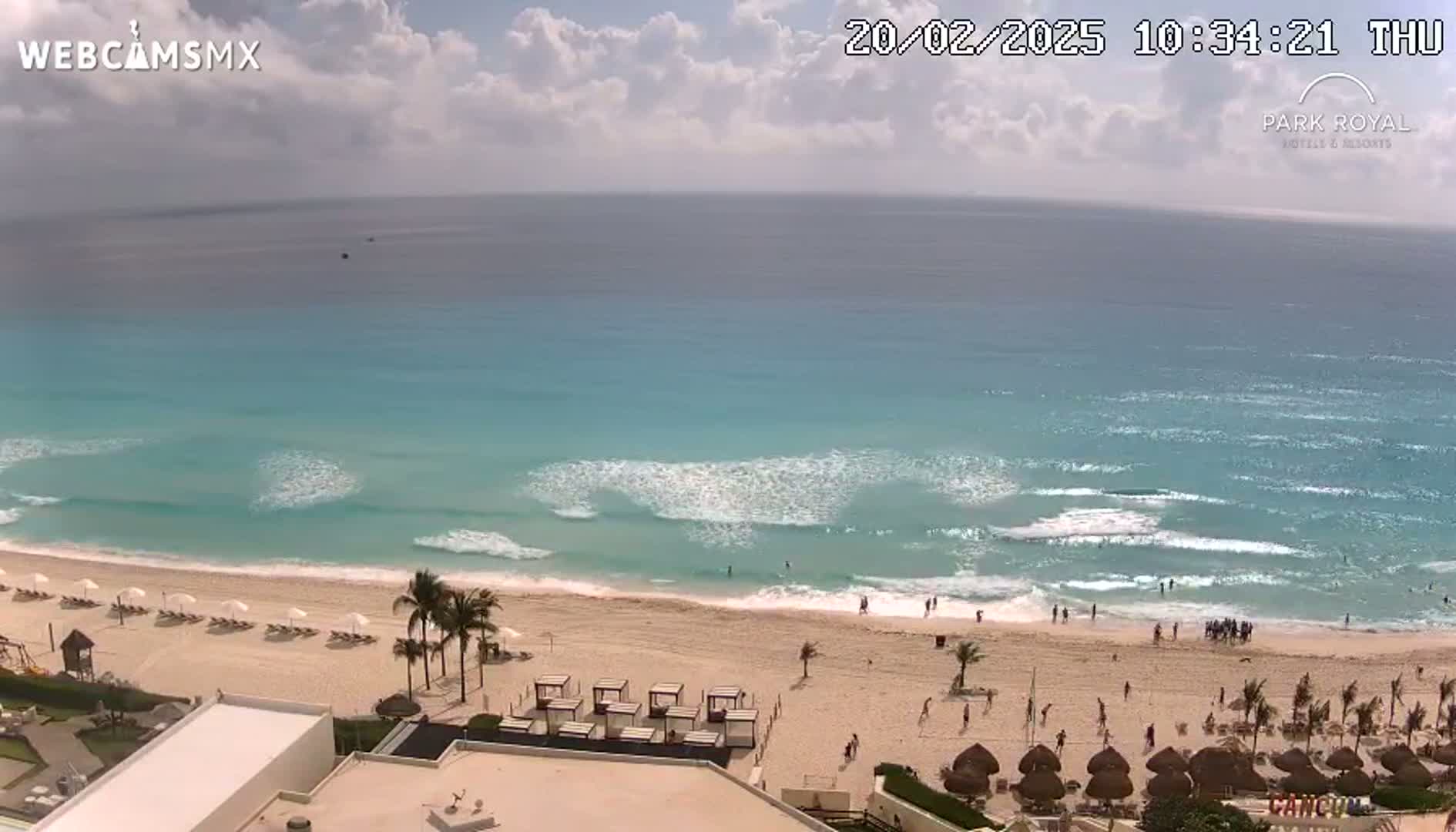 A mostly sunny day shows a long stretch of sandy beach with light waves lapping the shore, with people relaxing and playing on the sand and in the water.