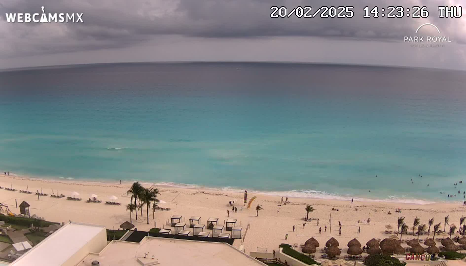 An aerial view of a beach with turquoise water under a partly cloudy sky shows people swimming and relaxing on the sand.